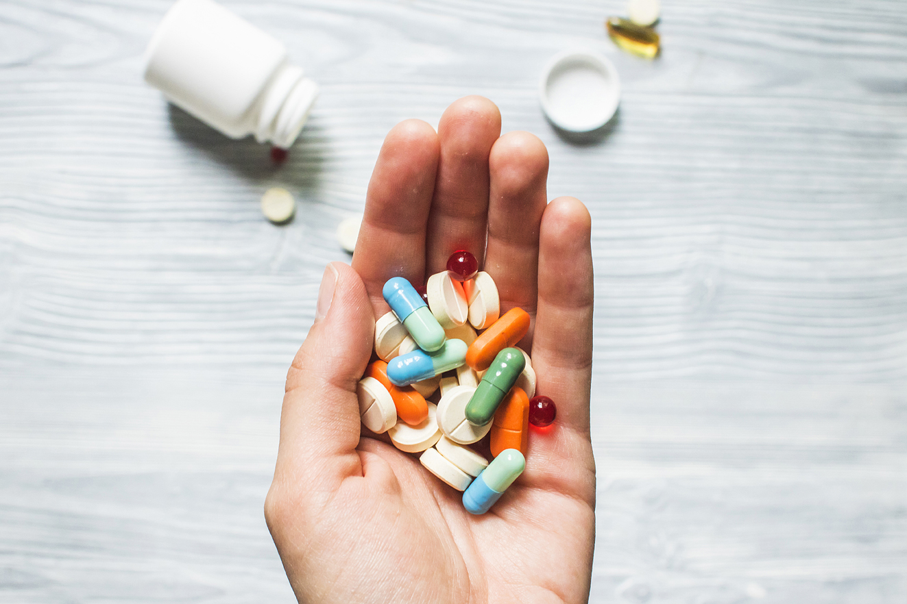 The palm of a hand filled with various pills and capsules against a white wood-grain background with an open pill bottle and medicine spilled out.