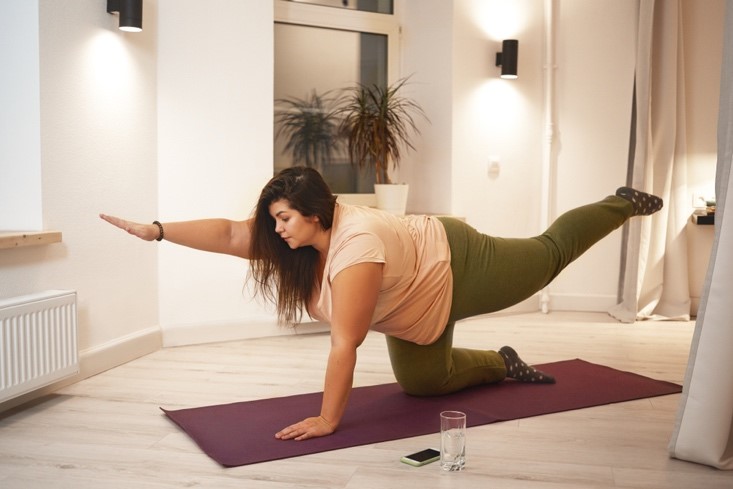 An overweight female in workout clothes practices yoga on a yoga mat.